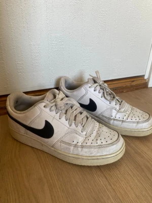 Vita Nike sneakers med svart swoosh - Klassiska vita Nike sneakers med svart swoosh på sidorna. Modellen har lågt skaft, perforerad tå och vit sula. Skorna är i läder och har vita skosnören. Perfekta för en avslappnad streetstil.