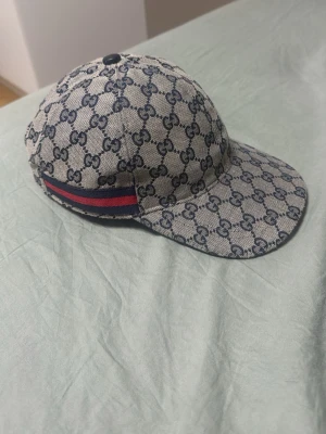 Gg keps Grey/Navy - Snygg keps från Gucci i beige och marinblått med klassiskt GG-monogram över hela kepsen. På sidan finns en ikonisk röd och marinblå rand. Kepsen har böjd skärm och är tillverkad i canvasmaterial.