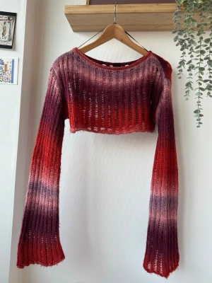 Stickad croppad tröja Urban Outfitters - Säljer en stickad croppad tröja från Urban Outfitters i röd, lila och rosa ombre. Tröjan har långa, vida ärmar. Mjukt och varmt material!