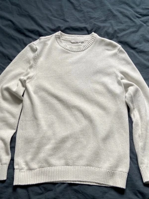 Vit stickad tröja med rund hals - En stilren vit stickad tröja med rund halsringning och ribbade muddar vid ärmslut och nederkant. Perfekt för lager-på-lager och enkel att matcha med olika outfits. Tröjan har en klassisk passform och är mjuk och bekväm.