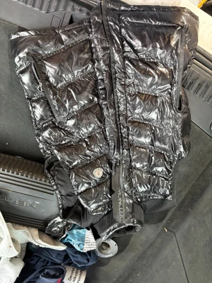 Svart glansig dunväst från Moncler - Snygg svart dunväst från Moncler med glansig finish och klassisk logotyp på bröstet. Västen har två stora fickor med dragkedja framtill och ribbade muddar nedtill. Insidan har ett serietidningsmönster och märkestaggar. Perfekt för lager-på-lager och streetwear.