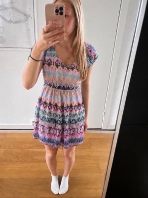 Färgglad kortklänning från Lindex - Kortklänning från Lindex med färgglatt mönster i rosa, blått, lila och gult. Klänningen har v-ringning, korta ärmar och paljett-detaljer vid halsen. Tillverkad i mjuk polyester, aldrig använd💕