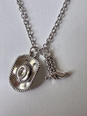 Silverfärgat halsband med cowboytema -  halsband i silverfärgad rostfritt metall med två hängen: en detaljerad cowboyhatt och en westerninspirerad stövel. Kedjan är av klassiska modellen ankarlänk och hängena har fina mönster och struktur. Perfekt accessoar för dig som gillar westernstil eller vill sticka ut med något annorlunda.