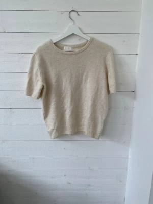 Beige stickad t-shirt från H&M - Säljer en beige stickad t-shirt från H&M med korta ärmar och rund halsringning. Den har lite smink fläckar som man kan se på bilden. Därav sänkt pris men kan diskuteras.