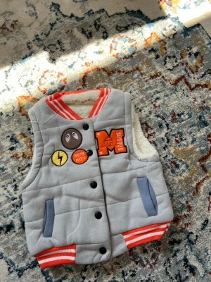 Grå dunväst med patches H.S.X.M.X - Grå dunväst med orange och vita detaljer från H.S.X.M.X. Västen har broderade patches med text och symboler, ribbade muddar i orange/vitt och mjukt vitt foder. Stängs med svarta knappar och har två fickor framtill. Perfekt för en cool och avslappnad stil.