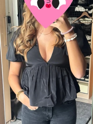 Nelly topp  - Trendig svart blus med djup v-ringning och korta puffärmar. Modellen har en lös passform med rynkad midja som ger en snygg siluett. Perfekt att styla med jeans eller kjol för en cool look.