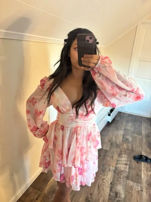 Blommig kortklänning med volang - Söt kortklänning med rosa och vita blommor, v-ringad och med långa puffärmar. Klänningen har volangdetaljer och markerad midja som ger en feminin siluett. Perfekt för sommarens alla tillfällen.