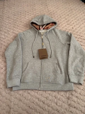 Burberry kofta - Snygg grå zip hoodie från Burberry med klassiskt rutigt foder i huvan och dragsko. Tillverkad i mjuk bomullsblandning, har ribbade muddar och fickor framtill. Perfekt för en avslappnad och stilren look.