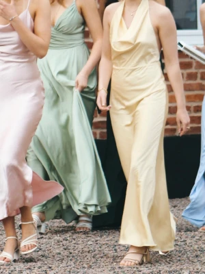 Ljusgul långklänning med halterneck - Elegant ljusgul långklänning i ett glansigt material med halterneck och draperad urringning. Klänningen har en figurnära passform och faller snyggt längs kroppen. Perfekt till balen eller för sommarens speciella tillfällen.