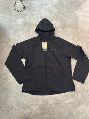 Svart nike Miler - Snygg svart vindjacka från Nike med huva och dragkedja framtill. Jackan har två sidofickor och diskret Nike-logga på bröstet. Tillverkad i lätt och tåligt material, perfekt för löpning eller andra utomhusaktiviteter. Modellen är enkel och sportig.