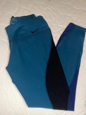 Blå Nike Dri-Fit leggings - Snygga blå leggings från Nike med Dri-Fit-teknologi. De har svarta och mörkblå paneler längs benen och en hög midja för extra komfort. Perfekta för träning eller chill. Materialet är stretchigt och andas bra.