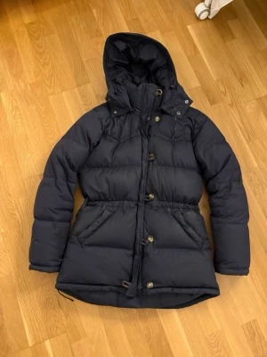 Mörkblå dunjacka med huva - Snygg mörkblå dunjacka med huva och knappar framtill. Jackan har dragsko i midjan för en mer figurnära passform och stora fickor på sidorna. Perfekt för kalla vinterdagar och har en klassisk, stilren look. Från boomerang 