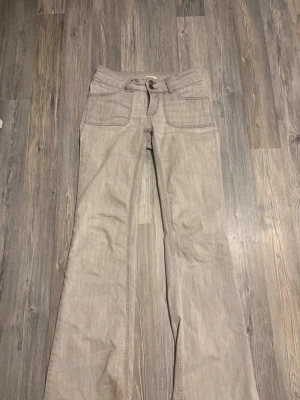 Ljusgrå bootcut lågmidjade jeans med fickor - Säljer ett par ljusgrå jeans med bootcut passform och stora framfickor. Jeansen har en klassisk knappstängning och är tillverkade i ett denimliknande material. Perfekt för dig som gillar en avslappnad och trendig stil.