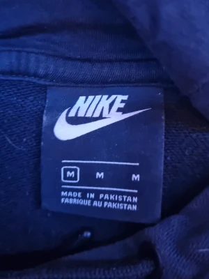 Svart hoodie från Nike med huva - Snygg svart hoodie från Nike med klassisk logga på bröstet. Hoodien har en huva med dragsko och är tillverkad i mjukt material som passar perfekt för en avslappnad stil. Perfekt för dig som gillar sportiga och bekväma plagg.