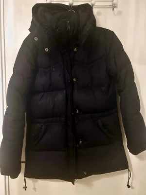 Mörkblå dunjacka med huva - Snygg mörkblå dunjacka från Boomerang med huva och dragsko nertill. Jackan har knappar och dragkedja framtill, två stora fickor, två innerfickor och en längre passform som håller dig varm under kalla dagar. Perfekt för dig som vill ha en stilren och funktionell vinterjacka.