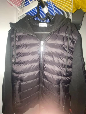 Svart Moncler cardigan  - Snygg svart dunväst från Moncler med huva och dragkedja framtill. Västen har quiltad framsida, mjuka ärmar och sidfickor med dragkedja. Moncler-logga på ärmen och cool streetstil. Perfekt för lager-på-lager och chill vardagslook.