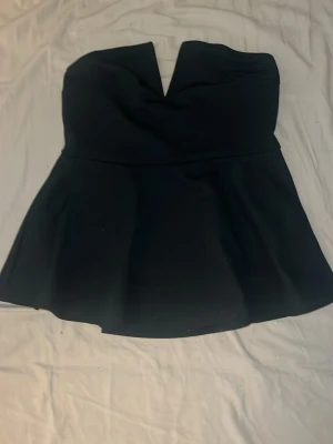 Svart peplum tubtopp med v-ringning - Snygg svart tubtopp med peplum-form och v-ringad detalj framtill. Toppen är axelbandslös och har en figurnära passning upptill som går ut i en volang nedtill. Perfekt för att skapa en trendig look.
