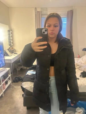 Svart lång dunjacka - Säljer en svart lång dunjacka med hög krage och quiltad design. Jackan har en rak passform och är perfekt för kalla vinterdagar. Den är tillverkad i ett tåligt material och har en stilren look som passar till jeans och sneakers.