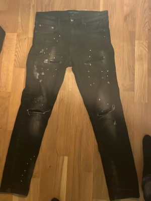 Svarta jeans supply and demand - Säljer ett par svarta slim fit jeans från supply n demand med coola slitningar och vita färgstänk på framsidan. Jeansen har klassisk femficksdesign och är tillverkade i stretchigt material för en bekväm passform. Perfekt för dig som gillar en edgy stil.