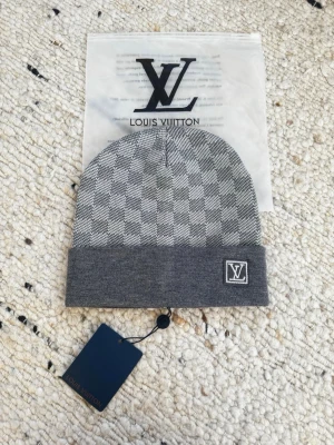 Grå L.V. mössa med rutmönster - Snygg grå mössa med klassiskt rutmönster och vikt kant. Designer-logga broderad framtill på kanten. Tillverkad i mjukt stickat material som känns lyxigt och bekvämt. Perfekt accessoar för att lyfta din stil. Kommer med prislapp och zipbag. Pris förhandlingsbart.
