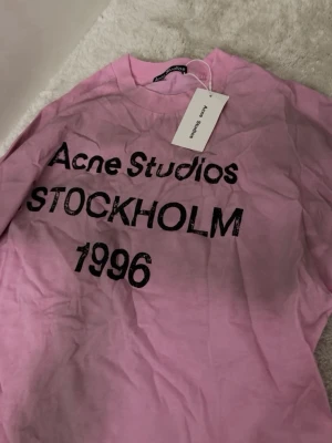 Rosa Tröja Acne Studios Logga - Säljer en rosa t-shirt från Acne Studios med svart text 'STOCKHOLM 1996' på framsidan. T-shirten är i 100% bomull och har klassisk passform med långa ärmar. Perfekt för dig som gillar streetwear och vill ha något från ett ikoniskt svenskt märke. Helt ny med pristagg och kvitto finns! Nypris 3500kr