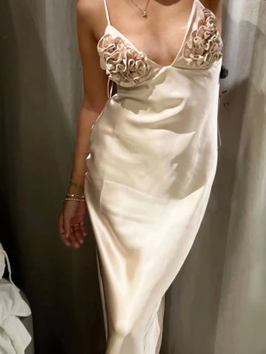 Champagnefärgad långklänning med volang - Elegant långklänning i champagnefärgad satin med smala axelband och djup v-ringning. Klänningen har dekorativa volanger över bysten och en slät, figurnära siluett. Perfekt för dig som vill ha en unik och stilren look.