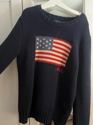Mörkblå stickad Ralph Lauren tröja  - Mörkblå stickad tröja från Polo Ralph Lauren med amerikansk flagga i rött, vitt och blått på bröstet samt RL-broderi. Tröjan har rund halsringning och ribbstickade muddar. Perfekt för dig som gillar klassisk och cool stil. Nypris ca 2000-3000