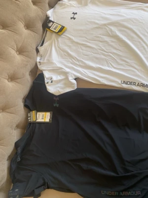 Under Armour set t-shirt & shorts - Säljer ett sportigt set från Under Armour med två t-shirts och två shorts. Ena t-shirten är vit och den andra svart, båda med logga på bröstet och tryck vid nederkanten. Shortsen kommer i svart och vitt, med elastisk midja och logga framtill. Perfekt för träning eller chill.
