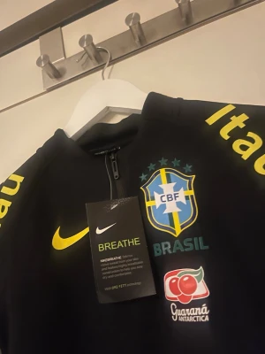 Svart Brasilien Nike träningströja - Svart långärmad träningströja från Nike med Brasilien CBF-emblem, gula och orange detaljer samt sponsorloggor. Stolek 120-130cm för barn 
