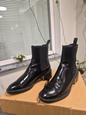 Zara svarta Chelsea boots i läder 36 - Snygga svarta Chelsea boots med klassisk design och elastiska sidpaneler. Skorna är tillverkade i blankt läder och har en låg klack samt dragflikar både fram och bak. Perfekta för dig som vill ha en stilren och tidlös look.