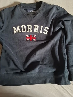 Mörkblå sweatshirt från Morris - Cool mörkblå sweatshirt från Morris med broderad Union Jack och vit text på bröstet. Tröjan har ribbade muddar och en liten patch på ärmen med Morris-logga och flagga. Perfekt för dig som gillar klassisk stil och snygga detaljer.
