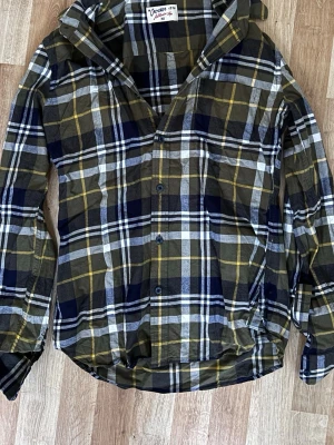Rutig skjorta från Crocker XS - Säljer en snygg rutig skjorta från Crocker i storlek XS. Skjortan har en mix av mörkblå, grön, vit och gul färg med klassiskt rutmönster, knappar framtill och långärmad design. Perfekt för dig som gillar en avslappnad och cool stil. Den passar mig utmärkt och jag är 170cm