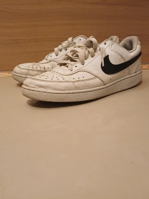 Vita Nike sneakers med svart swoosh - Klassiska vita sneakers från Nike med svart swoosh på sidan. Skorna har lågt skaft, perforerade detaljer på tån och ovandelen är i läder. Tidlös design med vita snören och slitstark sula. Perfekta för en streetwear-look.