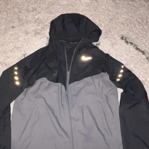 Nike svart och grå vindjacka - Nike vindjacka i svart och grått med huva och dragkedja. Jackan har reflekterande detaljer på axlar och rygg samt en vit Nike-logga på bröstet. Tillverkad i lätt och smidigt material, perfekt för blåsiga dagar. Sportig design med tvådelad färgblock.