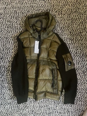 CP Company cardigan - Snygg olivgrön dunjacka från C.P. Company med stickade svarta ärmar och huva. Jackan har två framfickor med dragkedja och en ikonisk linsdetalj på vänster ärm. Perfekt för kalla vinterdagar och riktigt trendig look.