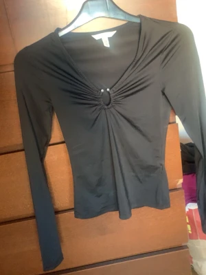 Svart långärmad topp med v-ringning - Snygg svart långärmad topp med v-ringning och rynkad detalj framtill som hålls ihop av en liten ring. Figurnära passform och mjukt, stretchigt material som känns bekvämt mot huden. Perfekt till jeans eller kjol för en trendig look.