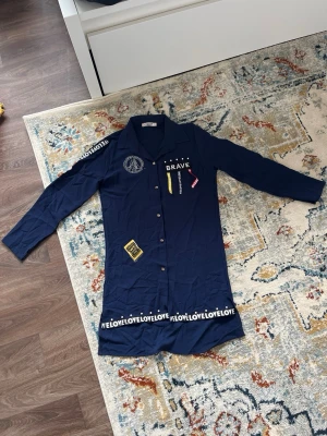 Marinblå skjortklänning med tryck - Marinblå skjortklänning från Allday med långa ärmar, knappar framtill och dekorativa tryck och banddetaljer i gult, vitt och rött. Texten 'LOVE' och 'BRAVE' samt broderad logga på bröstet ger en cool och ungdomlig stil.