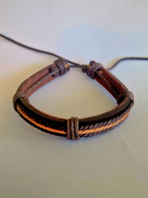 Brunt justerbart läderarmband - Snyggt armband i brunt läder med svarta och orangea detaljer. Armbandet har flera tunna remmar och är justerbart med knytning. Perfekt accessoar för en avslappnad och trendig stil.
