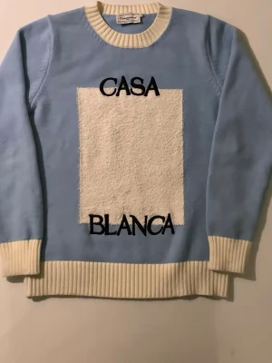 Blå stickad tröja från Casablanca - Säljer en snygg blå stickad tröja från Casablanca med kontrasterande vita muddar och krage. Framsidan har en stor vit, fluffig ruta och svart broderad text 'CASA BLANCA'. Perfekt för dig som gillar statement-plagg och vill sticka ut.