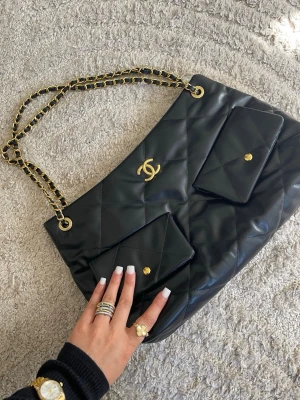 Svart quiltad axelväska med kedja - Snygg svart axelväska i quiltat konstläder med guldfärgad kedjerem. Väskan har en mjuk, puffig look och klassisk design som passar till många outfits. Perfekt för dig som vill ha en trendig och stilren accessoar.