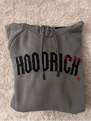 Grå HOODRICH hoodie med brodyr - Snygg grå hoodie från HOODRICH med svart broderad logga och röd detalj på bröstet. Klassisk huva med dragsko och stor magficka. Perfekt för en streetwear-look. Mjuk bomullsblandning och ribbade muddar.