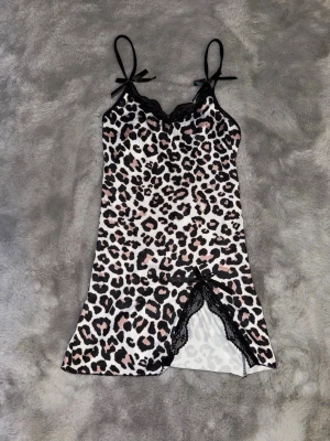 Leopardmönstrat nattlinne med spets - Snyggt nattlinne med leopardmönster i svart, vitt och ljusrosa. Tunna axelband med rosetter och spetsdetaljer vid urringningen och slitsen framtill. Kort modell i mjukt, stretchigt material som känns bekvämt mot huden. Liten i storlek. Helt ny.