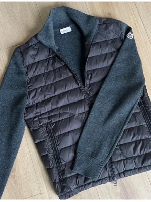 Moncler grå quiltad cardiganjacka - Snygg grå cardiganjacka från Moncler med quiltad front och stickade ärmar. Jackan har dragkedja framtill, två fickor med dragkedja och Moncler-logga på ärmen. Perfekt för lager-på-lager och stilrena outfits. Skriv för frågor.