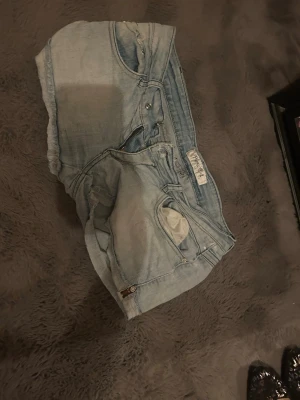Ljusblå jeansshorts från Levi's - Säljer ett par ljusblå jeansshorts från Levi's med klassisk femficksdesign och knappgylf. Shortsen har råa kanter nedtill för en avslappnad look och är tillverkade i denim. Perfekta för sommaren och passar till det mesta.