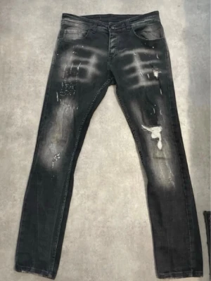 Svarta distressed jeans med knappar - Säljer ett par svarta jeans med slitna detaljer och blekta partier. Jeansen har knappgylf och klassisk femficksmodell. Materialet är denim och passformen är smal. Perfekta för dig som gillar en edgy och trendig stil.