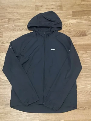 Svart Nike vindjacka med huva - Svart vindjacka från Nike med hel dragkedja och huva. Jackan har reflekterande detaljer på bröstet och ärmarna samt en minimalistisk design. Perfekt för blåsiga dagar och träning utomhus. Tillverkad i lätt och smidigt material.