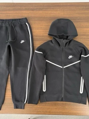 Svart Nike träningsset med reflex - Snyggt svart träningsset från Nike med matchande hoodie och byxor. Setet har vita reflexdetaljer längs ärmar, ben och bröst samt Nike-logga på både jacka och byxa. Perfekt för träning eller chill, med dragkedja på hoodien och resår i midjan på byxorna.