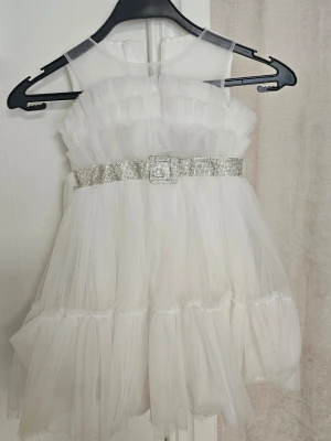Vit tyllklänning med glittrigt bälte - Supersöt vit kortklänning i tyll med volanger och ett glittrigt bälte med strassdetaljer i midjan. Klänningen är ärmlös och har dragkedja i ryggen. Perfekt för speciella tillfällen när du vill sticka ut lite extra.