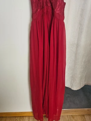 Röd långklänning med spets från ICHI - Elegant röd långklänning från ICHI med vacker spets upptill och ett flytande, lätt tyg i kjolen. Klänningen har ärmlös design och markerad midja, perfekt för speciella tillfällen.