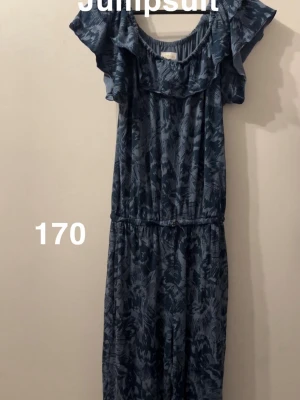 Mörkblå mönstrad jumpsuit med volang - Snygg mörkblå jumpsuit med abstrakt mönster och volangdetaljer vid axlarna. Resår i midjan för skön passform och rund halsringning. Perfekt för dig som gillar en avslappnad men trendig stil.🌸Slapp resår vid kragen. Storlek 170 